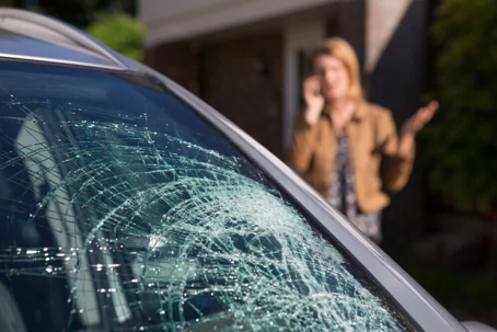 Blonde woman talking on cell phone behind car with broken windshield